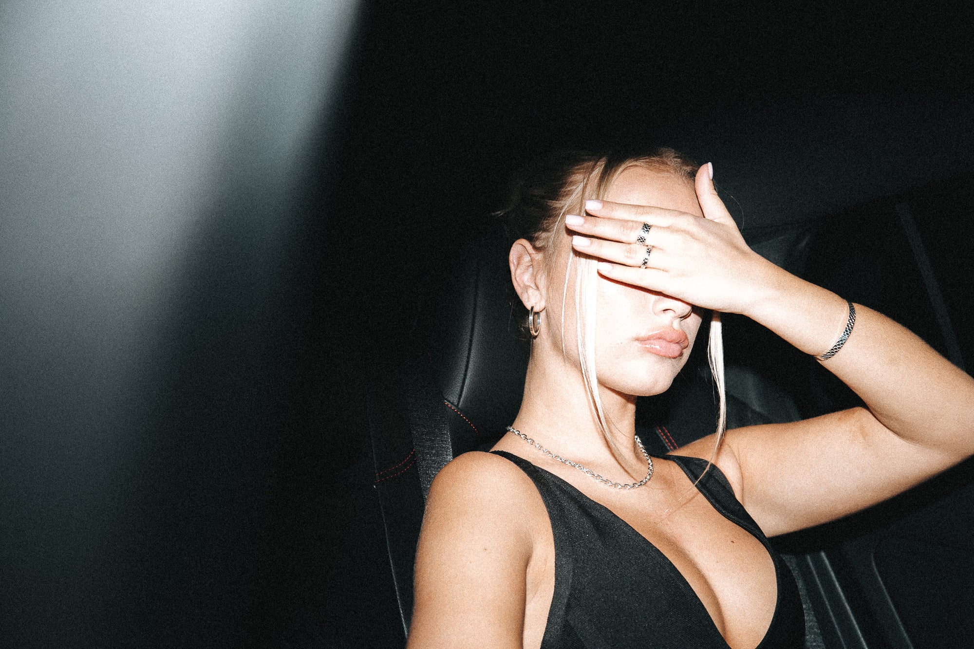 Woman in a black dress sitting in a car with one hand covering her face.